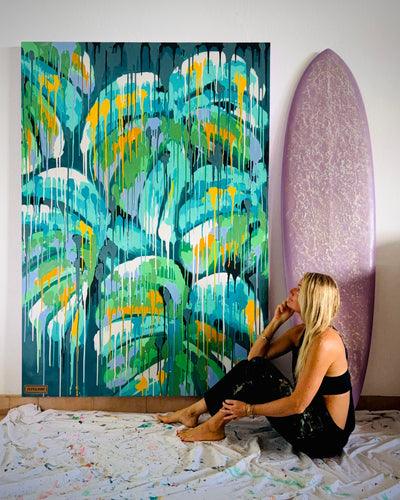 pepallama, tropical artist and surfer, sitting on the floor next to a large abstract original monstera painting and a surfboard.